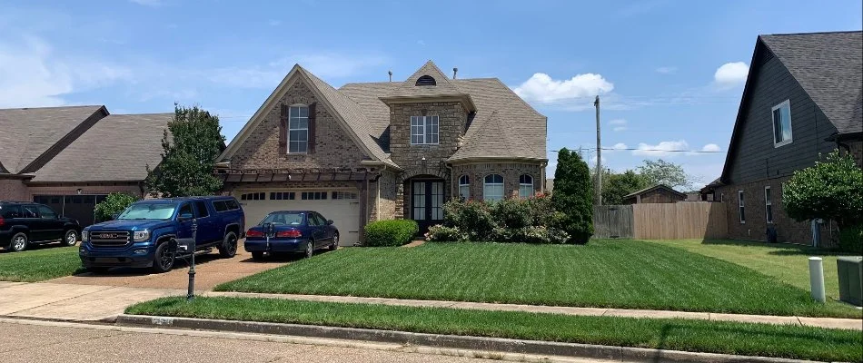 A stunning house with a stone and brick facade, featuring a well-manicured lawn and parked vehicles in a sunny neighborhood setting, with a clear blue sky and scattered clouds.